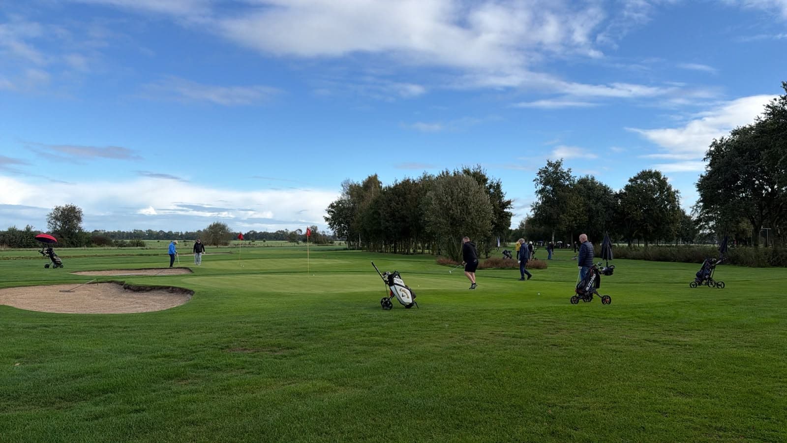 Golfers met trolleys op en rond een green met een bunker, onder een blauwe lucht met wolken.