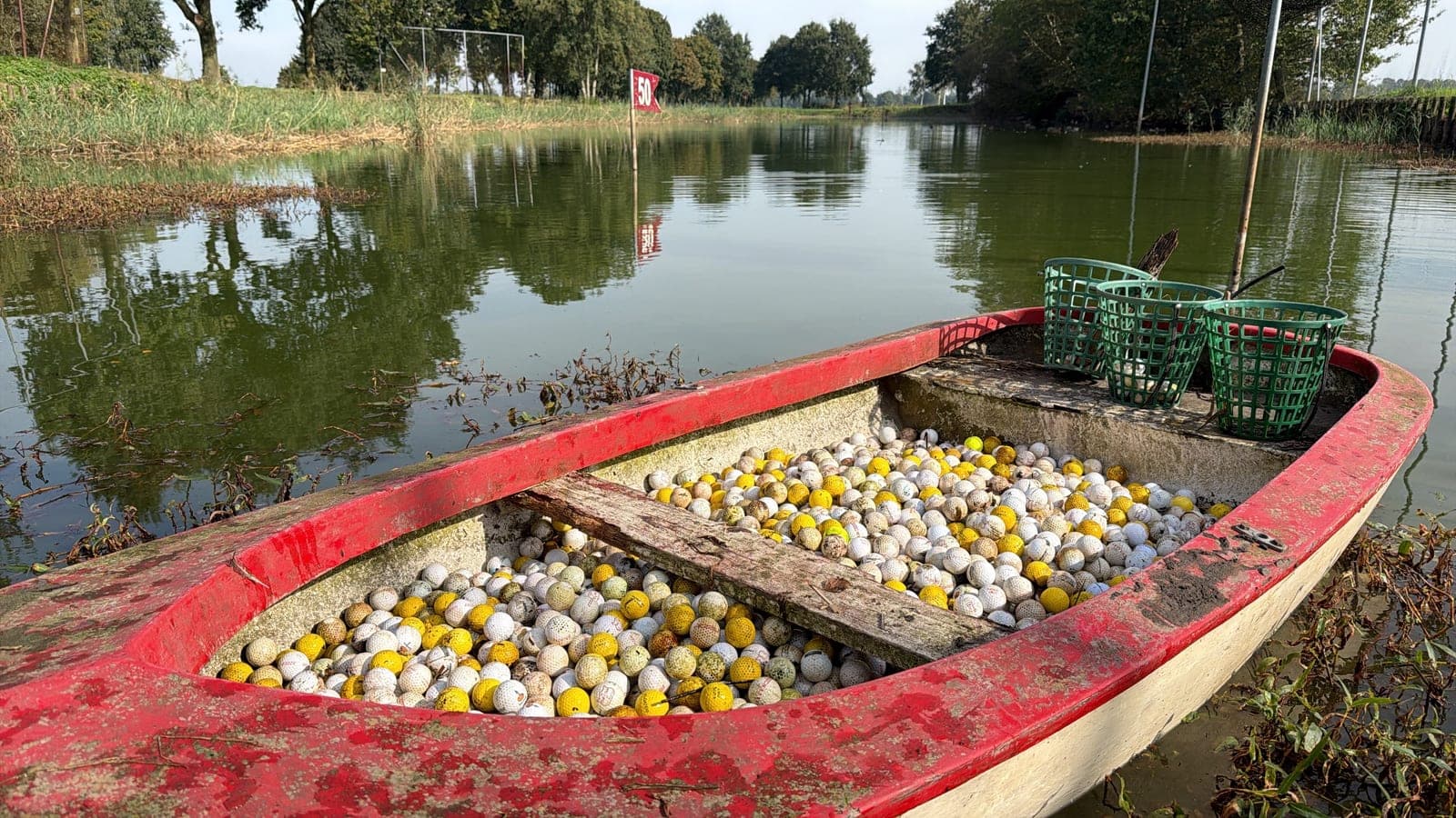 Een rood bootje vol witte en gele golfballen aan de rand van de vijver bij de driving range, met drie groene ballenmandjes erop.